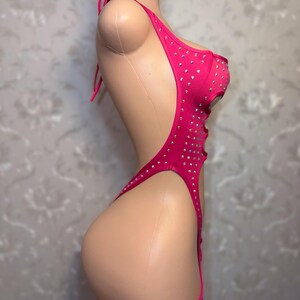 May include: A vibrant pink bodysuit adorned with rhinestones. This one-piece swimsuit has a halter neck, open sides, and a high-cut leg. The garment is displayed on a mannequin, highlighting its form-fitting design and decorative details.