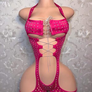May include: A vibrant pink bodysuit with a halter neck and lace-up detailing. The garment is adorned with many small, sparkling rhinestones. The design includes cut-out sections and attached shorts.