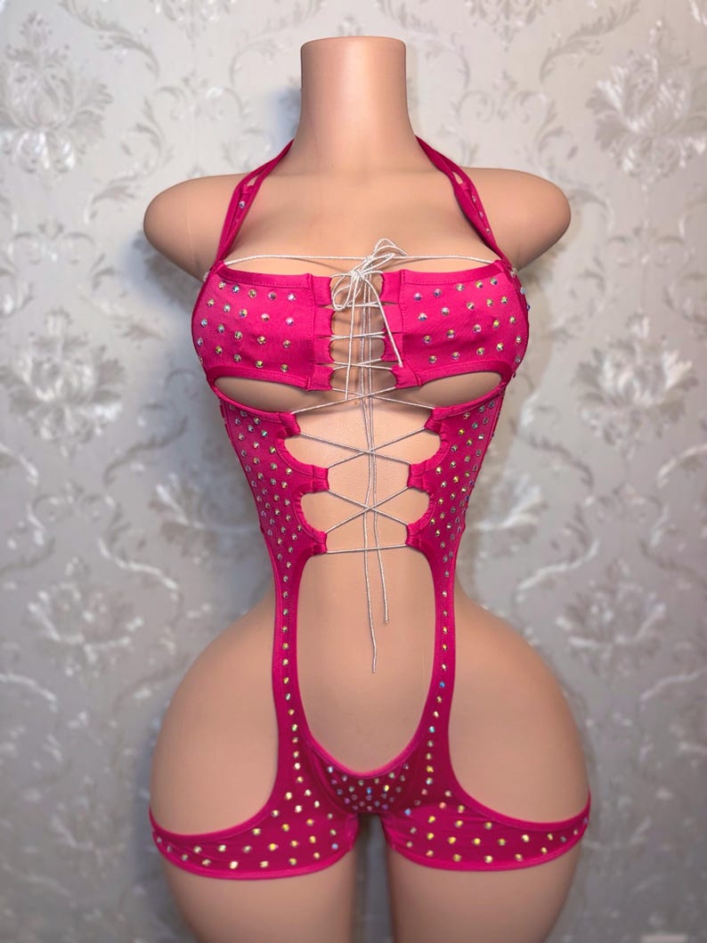 May include: A vibrant pink bodysuit with a halter neck and lace-up detailing. The garment is adorned with many small, sparkling rhinestones. The design includes cut-out sections and attached shorts.