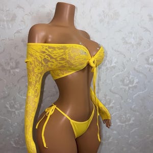 May include: A vibrant yellow lingerie set comprising an off-the-shoulder top with long sleeves and a bikini bottom. The top is crafted from lace and adorned with rhinestones. The bikini bottom features tie fastenings at the sides.