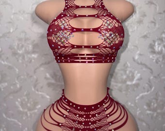 May include: A two-piece set consisting of a dark red fishnet crop top and matching high-waisted bottoms. The top features horizontal cutouts and iridescent rhinestones. The bottoms have side cutouts and are also embellished with rhinestones.