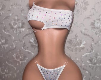 May include: White two-piece bikini set with iridescent rhinestone embellishments. The top is a one-shoulder design with a cutout, and the bottoms are a thong style. The set is displayed on a mannequin.