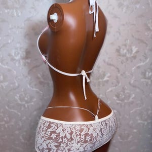 Peut inclure: Ensemble de lingerie en dentelle blanche sur mannequin. L'ensemble comprend un haut triangle &agrave; liens, un string et une mini-jupe avec un n&oelig;ud rose. La dentelle pr&eacute;sente un motif floral.