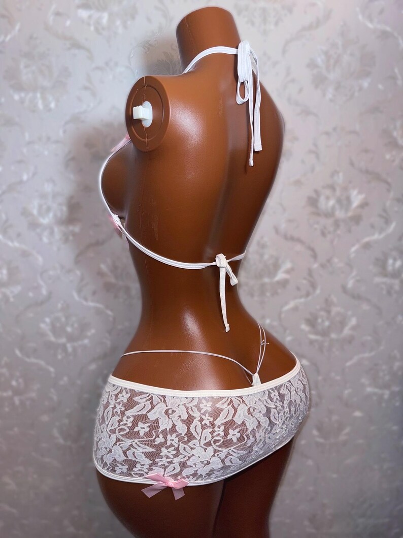 Peut inclure: Ensemble de lingerie en dentelle blanche sur mannequin. L'ensemble comprend un haut triangle &agrave; liens, un string et une mini-jupe avec un n&oelig;ud rose. La dentelle pr&eacute;sente un motif floral.