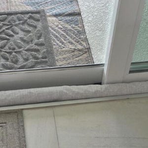 May include: Extra Long Door Draft Stopper #3 Solid Light Gray Fabric. A close-up of a light gray fabric door draft stopper on a white tile floor. The draft stopper is placed under a sliding glass door. A welcome mat with the word "HOME" is in the foreground.