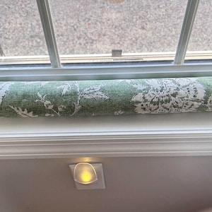 May include: A green and white floral patterned draft stopper, positioned beneath a window. The cylindrical stopper is made of fabric and rests on a white windowsill. The window frame is white, and the background shows an outdoor scene.