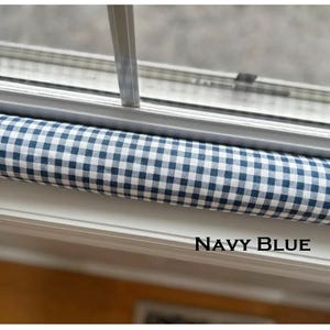 May include: A navy blue and white gingham patterned draft stopper, positioned beneath a window. The text "NAVY BLUE" is displayed in a black font at the bottom of the image. The stopper is cylindrical and appears to be made of fabric.