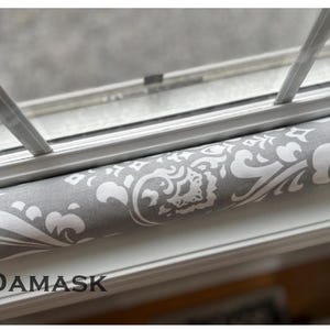 May include: A gray and white damask patterned window draft stopper. The fabric is wrapped around a cylindrical form, designed to fit snugly against a window frame. The word "DAMASK" is printed in black at the bottom.