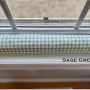 May include: Sage green and white gingham fabric window draft stopper. The fabric is a light green and white checkered pattern. The text "SAGE GREEN" is printed in black below the stopper.