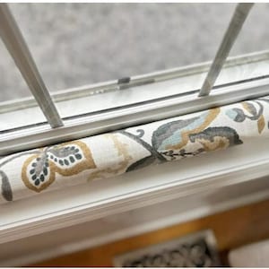 May include: A floral patterned window draft stopper in cream, gray, and gold tones. The fabric tube is positioned along the window frame, designed to block drafts and improve insulation. The design features botanical elements.