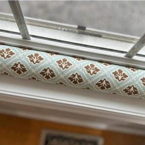 May include: A cylindrical draft stopper with a repeating pattern of brown floral shapes on a light blue background. The stopper is positioned beneath a window frame, offering a decorative and functional solution for sealing gaps.