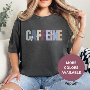 Puede incluir: Una camiseta gris oscuro con el texto "CAFFEINE" en letras de colores. El texto está en una fuente de estilo vintage. La camiseta la lleva puesta una persona. La imagen también incluye un círculo rosa con el texto "MÁS COLORES DISPONIBLES" y la palabra "Pepper" en texto blanco.