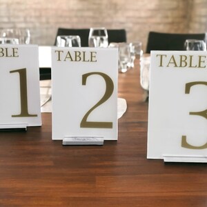 Op de afbeelding: Drie witte tafelnummerborden met gouden letters. De borden tonen "TABLE 1", "TABLE 2" en "TABLE 3".