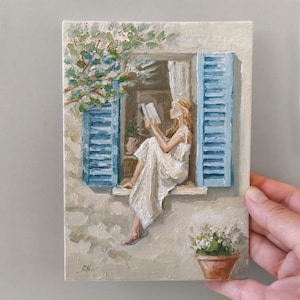 Peut inclure: Une peinture à l'huile sur toile représentant une personne en robe blanche assise dans un cadre de fenêtre aux volets bleus, lisant un livre. Du feuillage vert surplombe la fenêtre et un pot en terre cuite avec des fleurs blanches est en bas.