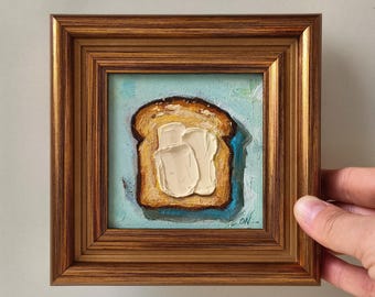 Puede incluir: Una pequeña pintura al óleo cuadrada en un marco de madera oscura que muestra una rebanada de pan tostado dorado con mantequilla blanca espesa y texturizada. El fondo es de color azul claro y las iniciales del artista ON se ven abajo a la derecha.