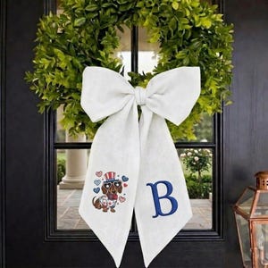 May include: A white bow with long tails hangs on a black door. The bow has a dachshund wearing sunglasses and a patriotic hat embroidered on one tail. The letter "B" is embroidered on the other tail. A green wreath is behind the bow.