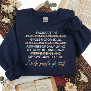 May include: Navy blue sweatshirt with colorful text: "I FACILITATE THE DEVELOPMENT OF FINE AND GROSS MOTOR SKILLS, SENSORY INTEGRATION, AND ACTIVITIES OF DAILY LIVING TO PROMOTE FUNCTIONAL INDEPENDENCE AND IMPROVE QUALITY OF LIFE. I help people do stuff."