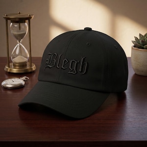 May include: Black baseball cap with the word "Blegh" embroidered in a gothic font. The cap has a curved brim and a button on top. The hat is sitting on a wooden surface.