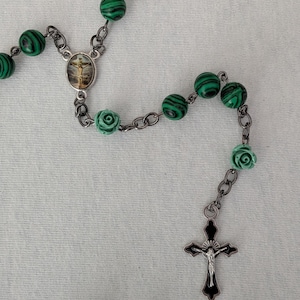 May include: A rosary with green and black patterned beads, silver-tone chain links, and a crucifix pendant. The rosary also features small, light green rose-shaped beads and a central medallion with a religious image.