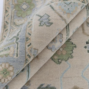 May include: Beige area rug with a detailed floral pattern in shades of blue and green. The rug is folded, showcasing the texture and design. The rug's border features a repeating geometric pattern.