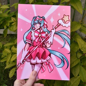 Può includere: Illustrazione rosa di un personaggio anime con lunghi capelli turchesi, che indossa un vestito rosa e tiene in mano un bastone sormontato da una stella. Lo sfondo è rosa con raggi di stelle e diamanti bianchi. L'opera è tenuta in mano.