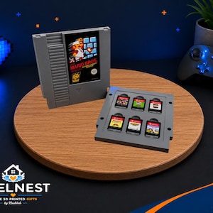 Puede incluir: Un cartucho gris impreso en 3D de Nintendo Entertainment System (NES) y un soporte de cartucho gris con seis cartuchos de juego. El cartucho de NES presenta la etiqueta del juego "Mario Bros.". Los artículos se muestran sobre una superficie de madera con un corazón pixelado azul y un mando en el fondo.