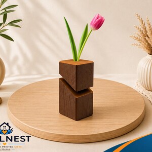 Op de afbeelding: Een moderne, geometrische houten vaas met een roze tulp. De vaas is gemaakt van twee gestapelde, schuine blokken donkerbruin hout. Andere decoratieve elementen zijn een witte vaas met groen, een kleine vetplant en een brandende kaars.