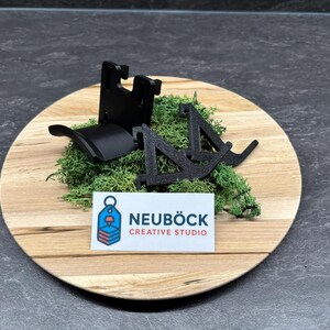 Puede incluir: Objetos negros impresos en 3D sobre una placa de madera con musgo verde. Los objetos parecen ser componentes de un soporte. También hay una tarjeta de visita con el texto "NEUBÖCK CREATIVE STUDIO".