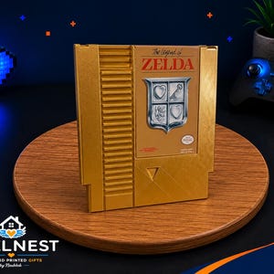 Könnte beinhalten: Eine goldfarbene Nachbildung einer klassischen Videospielkassette für die NES-Konsole. Das Etikett zeigt den Text The Legend of ZELDA in Rot sowie ein silbernes Schildelement mit Herz- und Schlüsselsymbol. Die Kassette hat eine texturierte Seitengrifffläche.