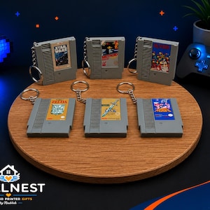 Könnte beinhalten: Graue NES-Kassetten-Schlüsselanhänger mit Spieletiteln wie Star Wars und Zelda. Die Schlüsselanhänger sind auf einer Holzoberfläche mit einem blauen Pixel-Herz und einem Gamecontroller im Hintergrund angeordnet. Das Pixel Nest Logo ist sichtbar.