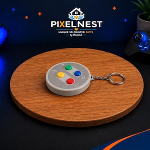 Può includere: Un portachiavi grigio a forma di controller di videogiochi classico, con pulsanti colorati. Il portachiavi è attaccato a un anello d'argento e poggia su una superficie rotonda di legno. L'immagine include anche un cuore blu brillante e un controller di gioco.
