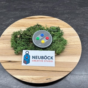 Op de afbeelding: Een grijze sleutelhanger in de vorm van een klassieke videogamecontroller met kleurrijke knoppen. De sleutelhanger ligt op een bedje van groen mos op een ronde houten plank. Een visitekaartje met "NEUBÖCK CREATIVE STUDIO" is ook aanwezig.