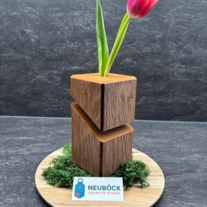 Könnte beinhalten: Eine braune Holzvase mit strukturiertem Holzmaserungsdesign, die eine rosa Tulpe mit grünen Blättern hält. Die Vase steht auf einem Holzbrett mit grünem Moos. Die Visitenkarte zeigt "NEUBÖCK CREATIVE STUDIO."