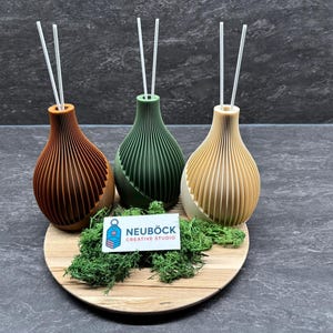 Puede incluir: Tres difusores de caña decorativos en tonos marrón, verde y beige, cada uno con dos cañas blancas. Los difusores están sobre una bandeja de madera con musgo verde y una tarjeta de visita "NEUBÖCK CREATIVE STUDIO."