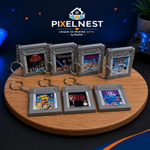 Pode incluir: Seis chaveiros cinzentos de cartuchos de Game Boy com arte de jogos clássicos, incluindo Star Wars e Donkey Kong, dispostos numa bandeja de madeira. O logotipo PixelNest é visível no fundo.