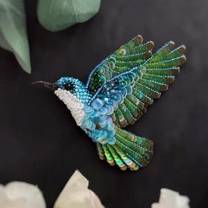 Könnte beinhalten: Handgefertigte Kolibri-Brosche mit einem detaillierten Design aus blauen, grünen und weißen Pailletten und Perlen. Der Vogel ist im Flug dargestellt, mit ausgebreiteten Flügeln, die die aufwendige Paillettenarbeit zeigen. Die Brosche ist vor einem dunklen Hintergrund platziert.