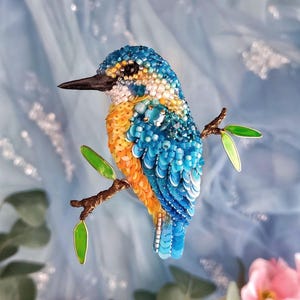 May include: A beaded kingfisher bird perched on a branch with green leaves. The bird is blue, orange, and white with a black eye. The background is a light blue fabric with pink flowers.