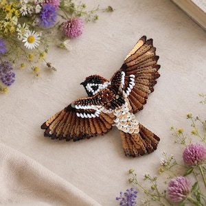 May include: A handcrafted bird brooch featuring intricate beadwork and sequins in shades of brown, white, and black. The bird is depicted in flight with wings fully extended, showcasing detailed plumage patterns created with layered sequins and beads.