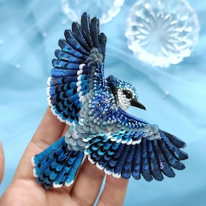 Può includere: Spilla artigianale a forma di ghiandaia azzurra con ali spiegate, dettagliata con paillettes e perline. L'uccello presenta sfumature di blu, bianco e grigio, creando un accessorio realistico e decorativo. La spilla è progettata per essere indossata come accessorio moda.