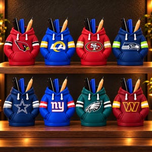 Puede incluir: Una colección de ocho organizadores de escritorio con forma de sudaderas con capucha en miniatura, cada uno con el logotipo de un equipo de la NFL diferente. Están dispuestos en dos filas y contienen bolígrafos y lápices.
