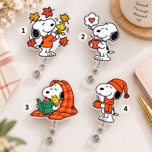 May include: Four acrylic badge reels featuring Snoopy and Woodstock. Design 1 shows Snoopy with autumn leaves. Design 2 shows Snoopy with a coffee mug and heart thought bubble. Design 3 shows Snoopy and Woodstock under a plaid blanket reading a book.