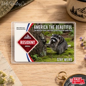 K&ouml;nnte beinhalten: Ein rechteckiger America the Beautiful National Parks and Federal Recreational Lands Pass in einer durchsichtigen Kunststoffh&uuml;lle. Die Karte zeigt einen Waschb&auml;ren an einem Baum, ein rotes rautenf&ouml;rmiges Resident Annual Etikett und den Text Stay Weird.