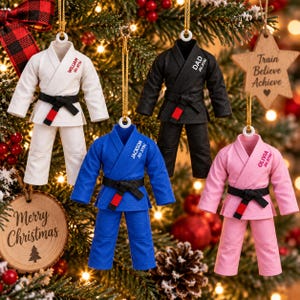 May include: Four miniature martial arts uniform ornaments in white, black, blue, and pink, each featuring a black belt with a red stripe. The uniforms are personalized with names and the text Jiu Jitsu. They hang from gold strings on a tree.
