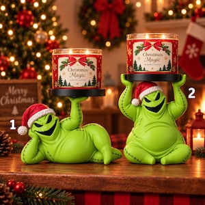 May include: Two green, stitched character candle holders wearing red and white Santa hats, each supporting a cylindrical glass jar candle labeled Christmas Magic with a red bow, green pine trees, and gold snowflake graphics on the front.