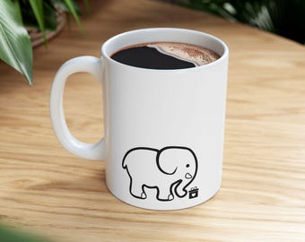 May include: White ceramic coffee mug with a black outline illustration of an elephant and a small gift box. The mug is filled with dark coffee. The mug has a curved handle and is sitting on a wooden surface.