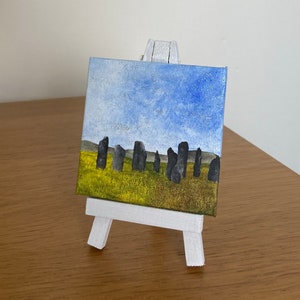 May include: A small painting on a white easel depicts a landscape with a row of dark grey stones in a field of green and yellow grass under a blue sky with white clouds.