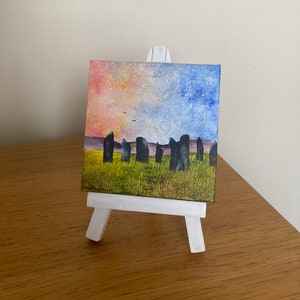May include: A small painting on canvas depicting a landscape with a row of dark grey standing stones in a field of green grass. The sky is painted in shades of pink, blue, and white with a few small birds flying in the sky.