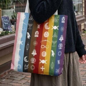 Op de afbeelding: Een rechthoekige canvas draagtas met een verticaal regenboogstrepenpatroon. Elke kleurstrook is versierd met witte lijnkunstsymbolen, waaronder manen, sterren, bomen, handen en geometrische vormen, gedragen aan twee witte stoffen schouderbanden.