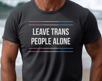 Peut inclure: Un t-shirt gris foncé avec le texte "LEAVE TRANS PEOPLE ALONE" en blanc. Le texte est centré sur le t-shirt et est entouré de trois lignes horizontales, deux roses et une bleue.