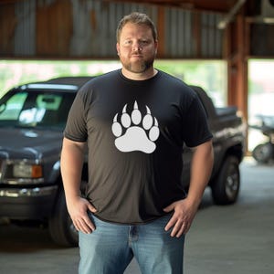 Puede incluir: Camiseta negra con una gran huella de pata de oso blanca. La camiseta es de cuello redondo y la lleva una persona. La persona lleva vaqueros azules y está de pie delante de una camioneta.